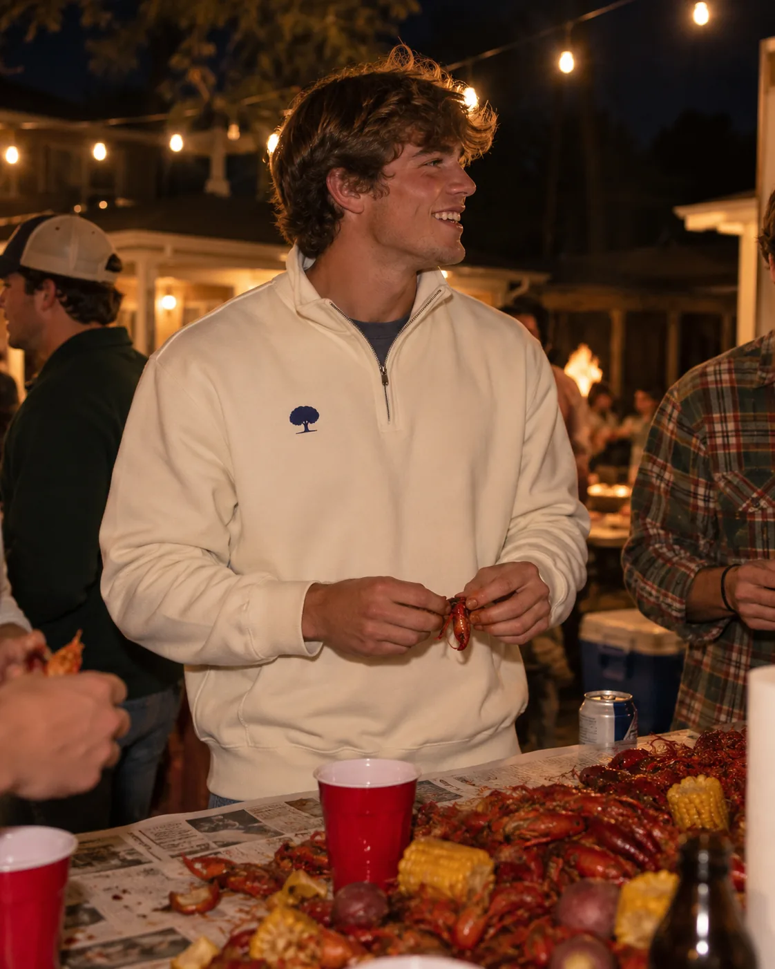 A young man at a nighttime crawfish boil under string lights wearing the cream Blue Oak Cream Quarter Zip